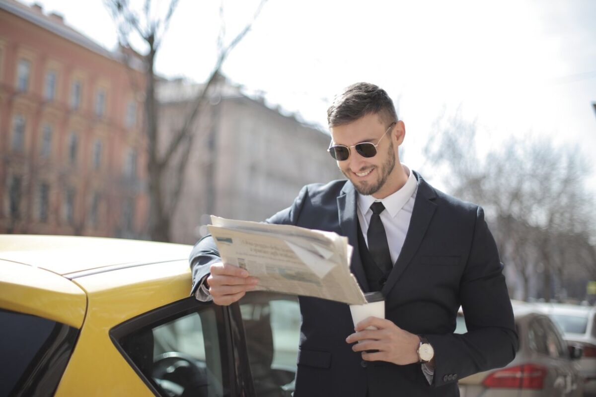 Offres A stylish businessman in a suit reads a newspaper outside by a car, holding a coffee cup.