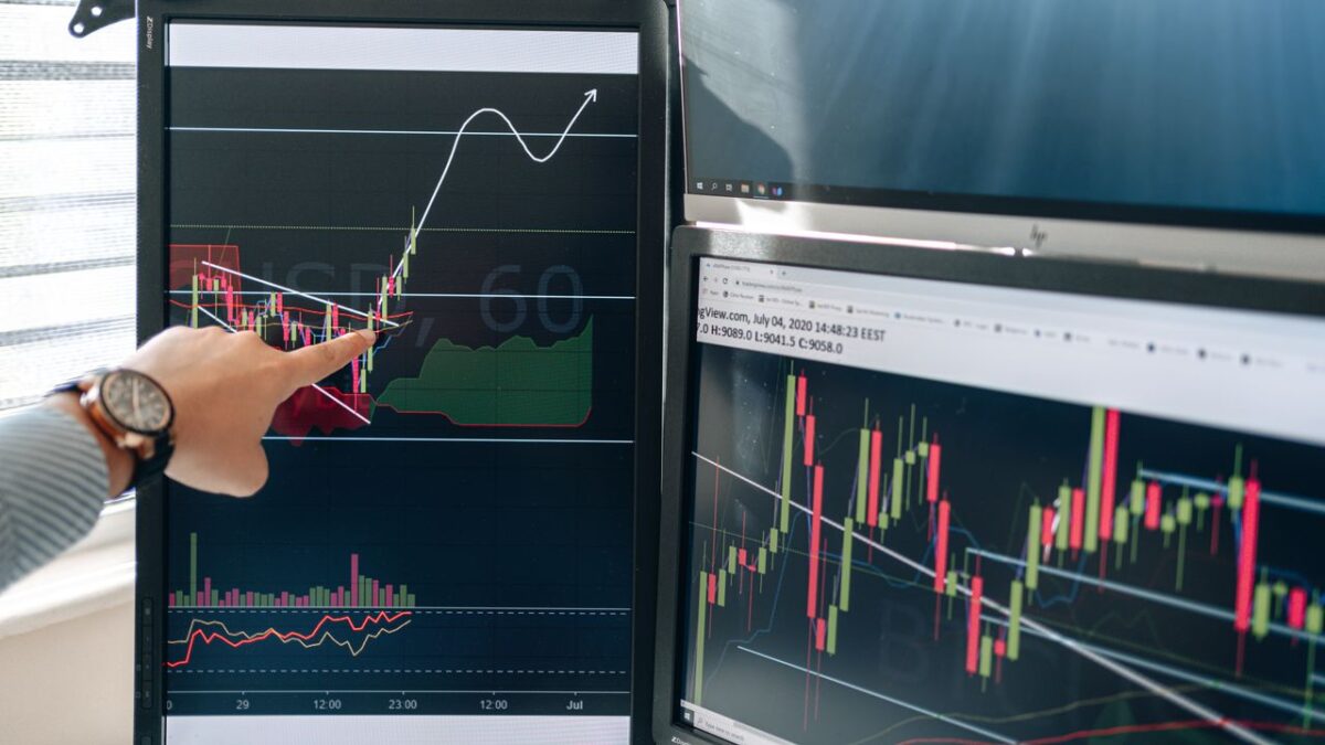 Offres Person pointing at stock market graphs on dual monitors in a modern workspace.